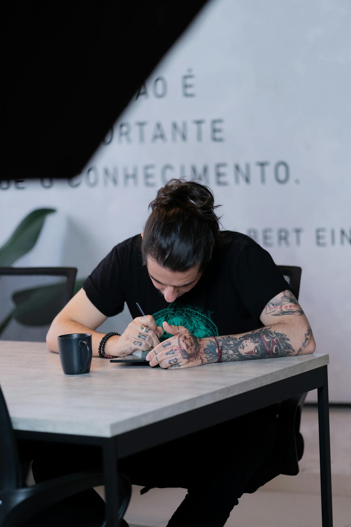 A tattooed person intensely writes at a desk in a modern workspace.